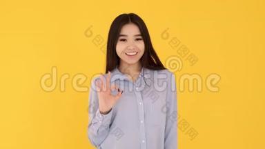 一个穿着<strong>蓝色</strong>衬衫的年轻女孩在<strong>黄色背景</strong>上微笑，并显示OK标志